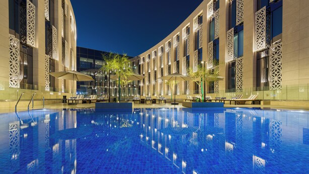 A swimming pool with lounge chairs and umbrellas, surrounded by a building with patterned windows and a tiled floor. - IntercityHotel Muscat (Muscat)