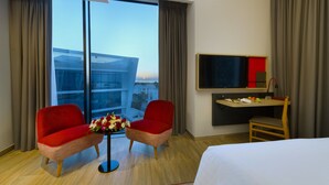 A hotel room with a large window, a bed, a desk with a TV, two red chairs, and a small table with flowers. - IntercityHotel Muscat (Muscat)