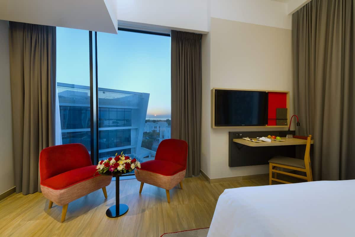 Ein Hotelzimmer mit großem Fenster, einem Bett, einem Schreibtisch mit Fernseher, zwei roten Sesseln und einem kleinen Tisch mit Blumen.