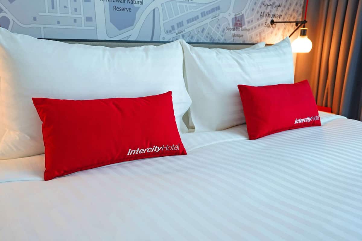 Ein Hotelbett mit weißer Bettwäsche und zwei roten Kissen mit dem Intercity Hotel Logo.