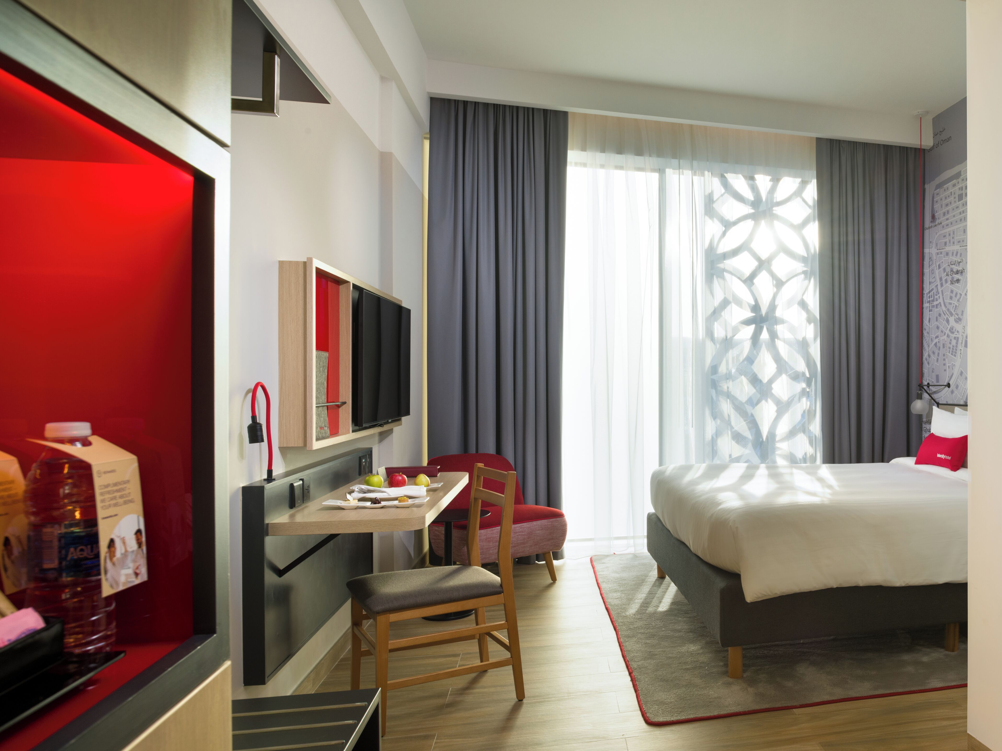 Une chambre d’hôtel avec un lit, un bureau avec une télévision, une chaise, un coussin rouge et une grande fenêtre avec des rideaux.