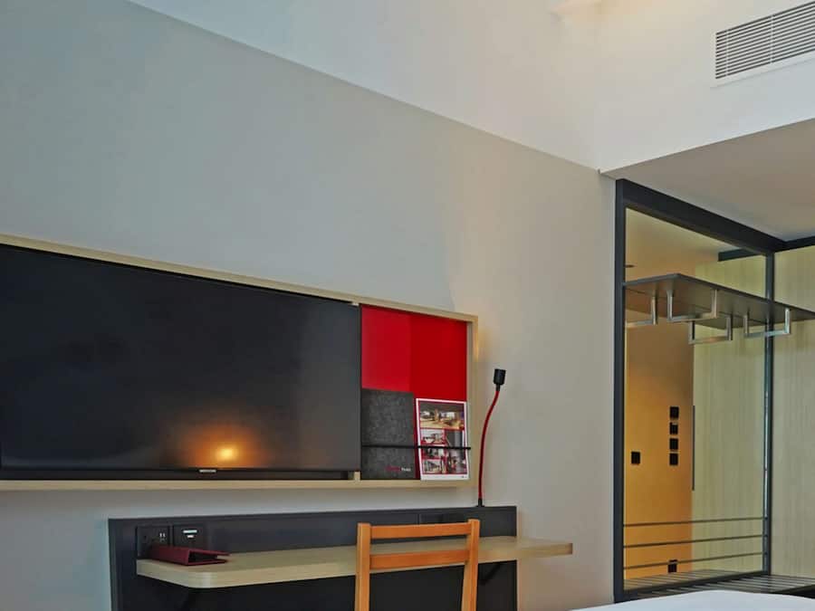Quarto de hotel moderno com uma grande janela, uma mesa com cadeira, uma estante de livros e um espelho.