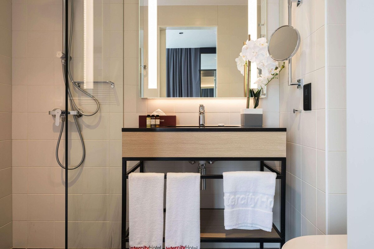 A modern bathroom with a shower, sink, and mirror. There are towels hanging on a rack and a vase with flowers on the counter.