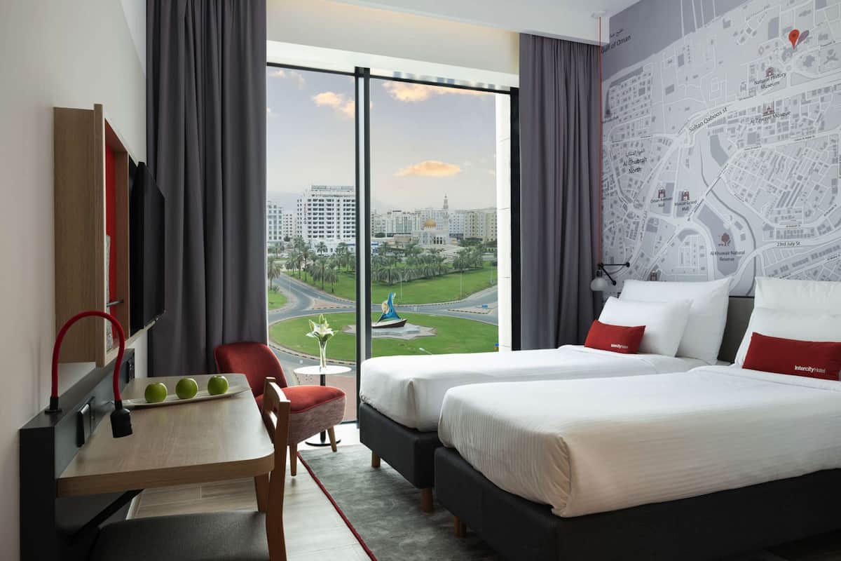 Ein Hotelzimmer mit zwei Betten, einem Schreibtisch mit Äpfeln, einem Sessel und einem großen Fenster mit Blick auf die Stadt.