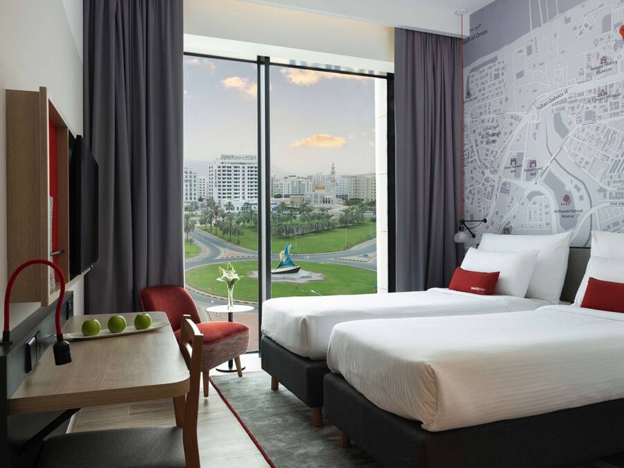 Quarto de hotel com duas camas, uma escrivaninha com maçãs, uma cadeira e uma janela grande com vista para a cidade.