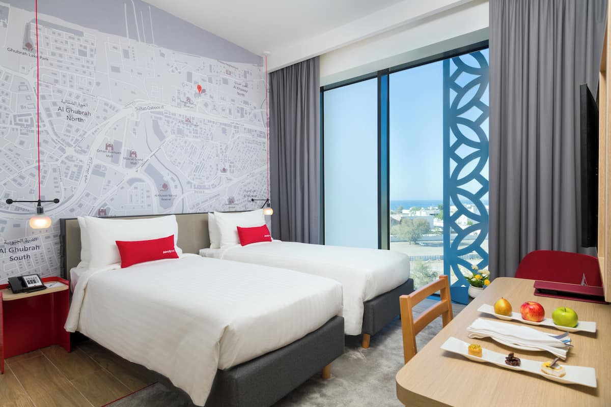Ein Hotelzimmer mit zwei Betten, einem Schreibtisch mit Früchten, einer Stadtansicht an der Wand und Blick auf die Stadt.