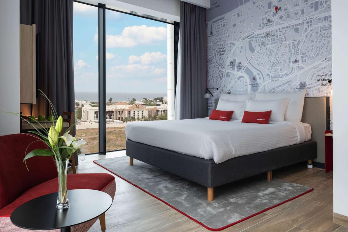 Ein modernes Hotelzimmer mit einem großen Bett, einem roten Sessel, einer Blumenvase und Blick auf eine Stadtlandschaft durch große Fenster.