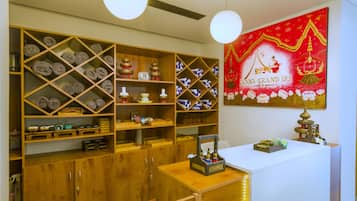 Un vestíbulo de hotel con estanterías de madera que exhiben toallas y objetos decorativos, un mostrador de recepción con un pequeño altar, y una gran pintura mural roja.