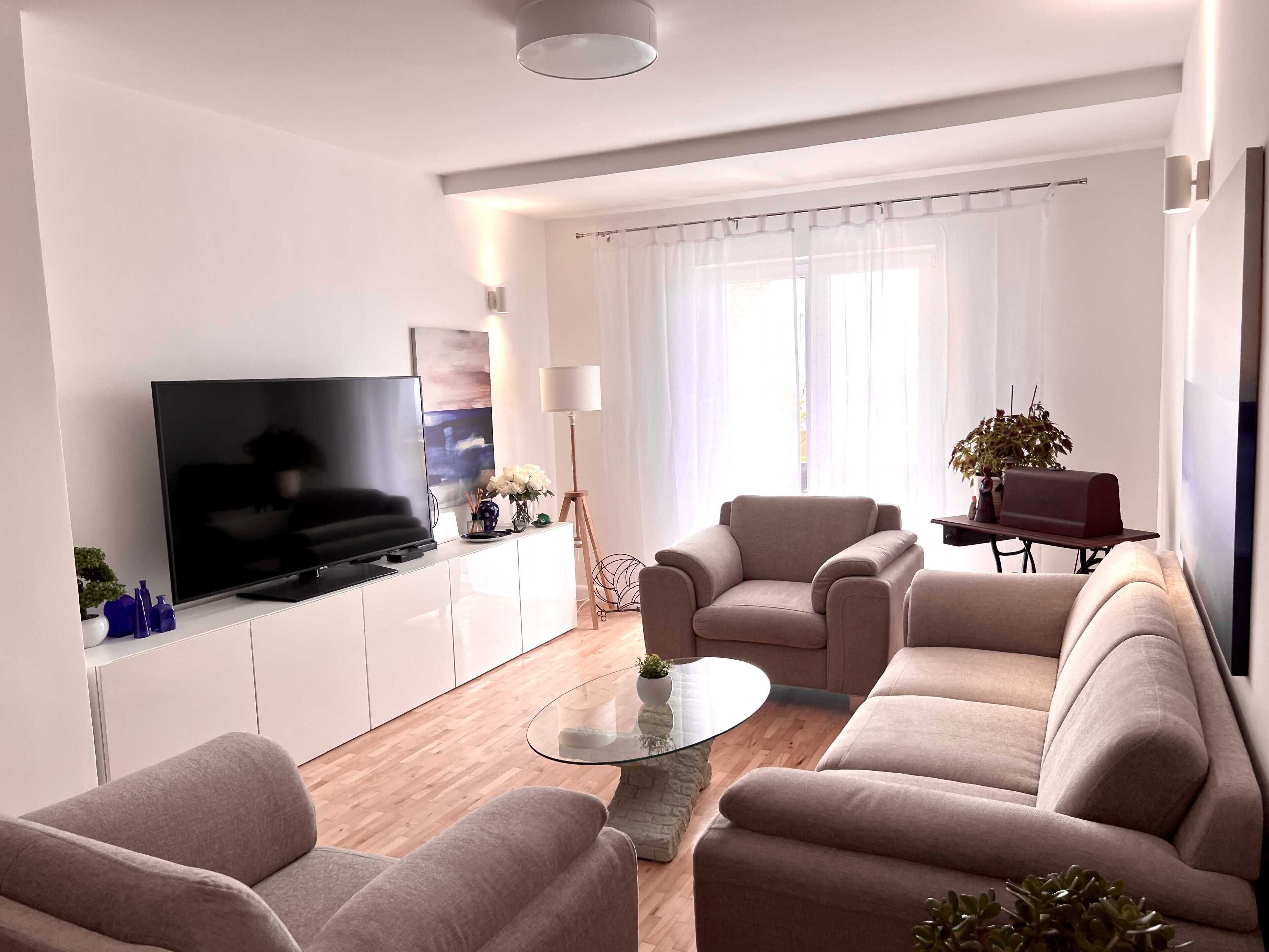 Una sala de estar moderna con un televisor de pantalla plana, sofás beige, una mesa de centro de vidrio y una planta en maceta.