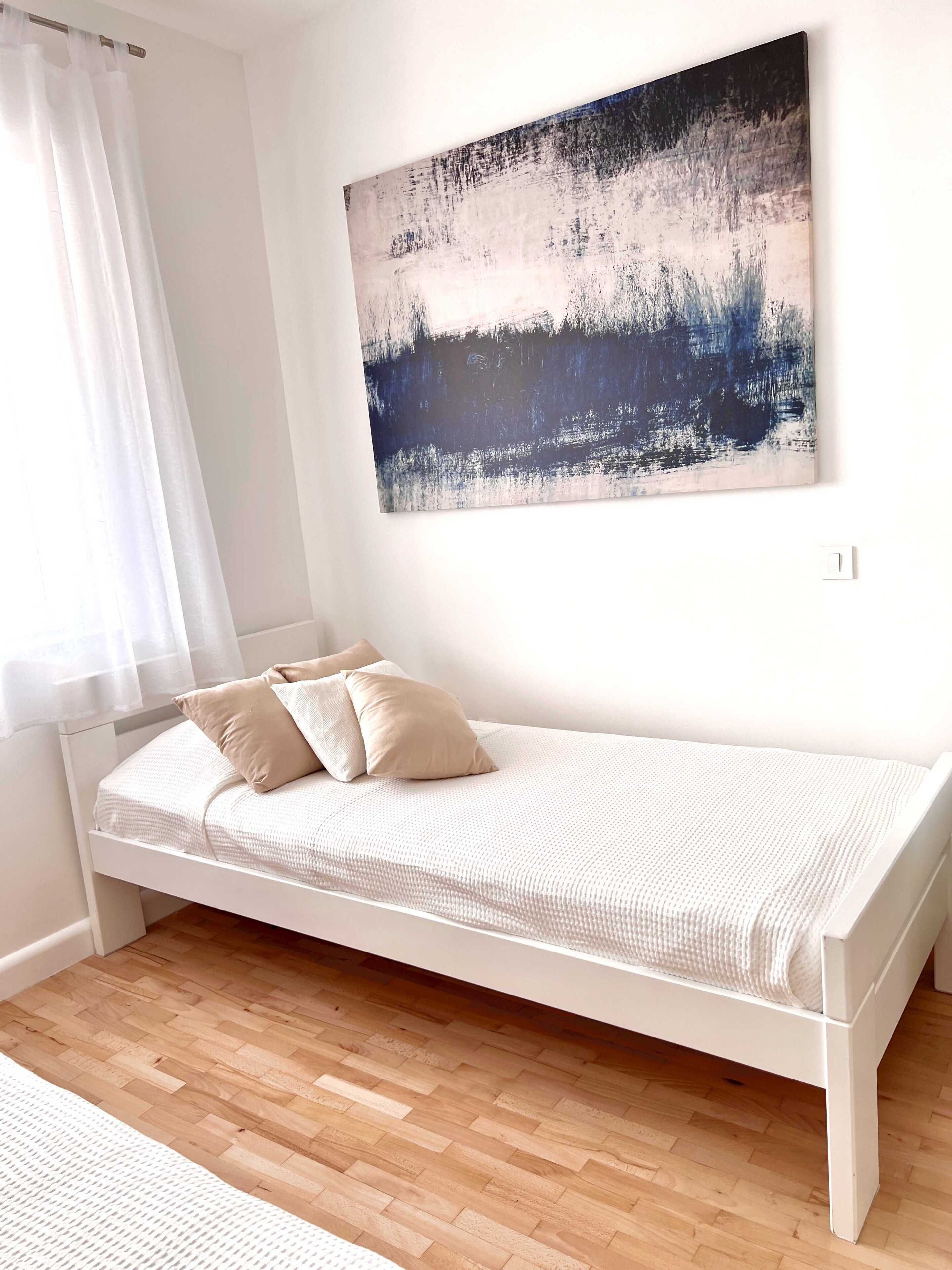 Un lit simple avec un cadre blanc et une literie beige, un mur blanc et une grande peinture abstraite accrochée au-dessus.
