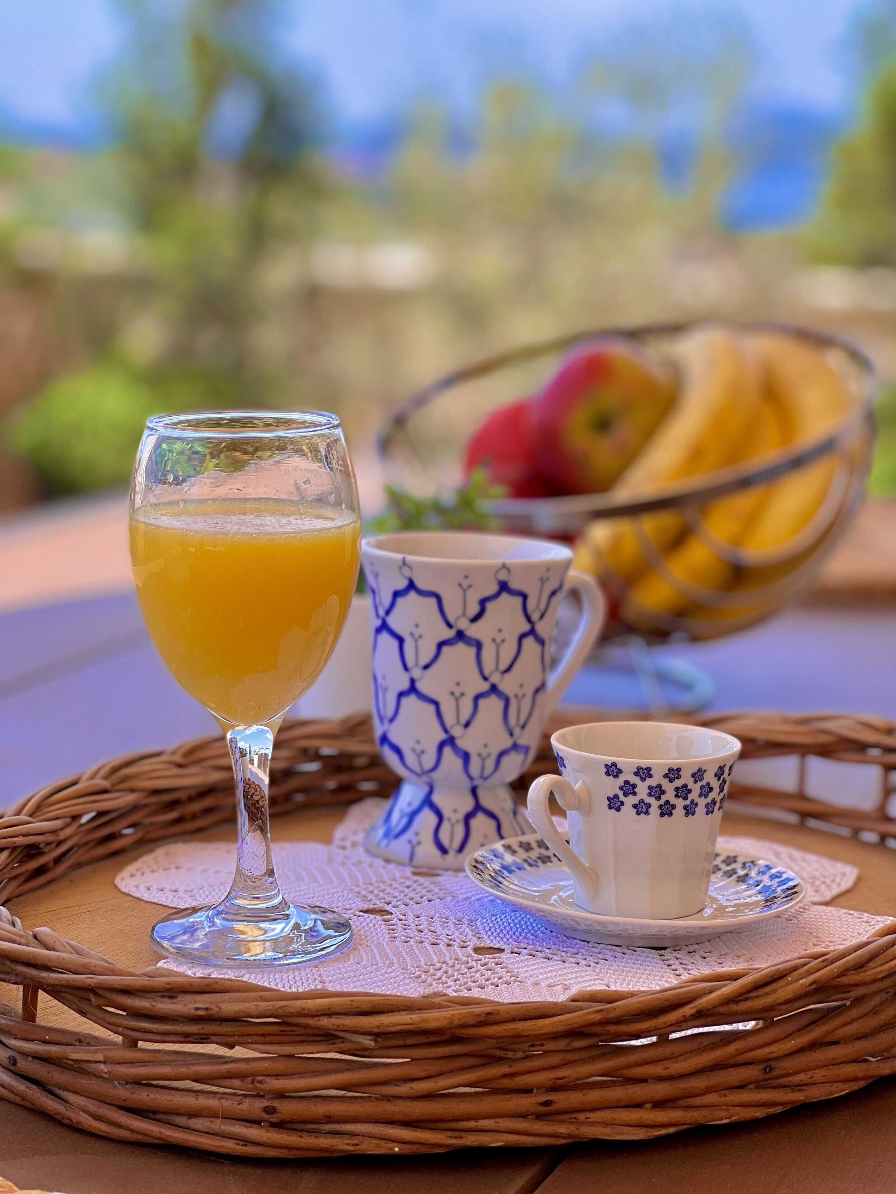 A wicker tray with a glass of orange juice, a blue and white patterned cup, and a small white cup with floral designs, all on a table with a lace doily.