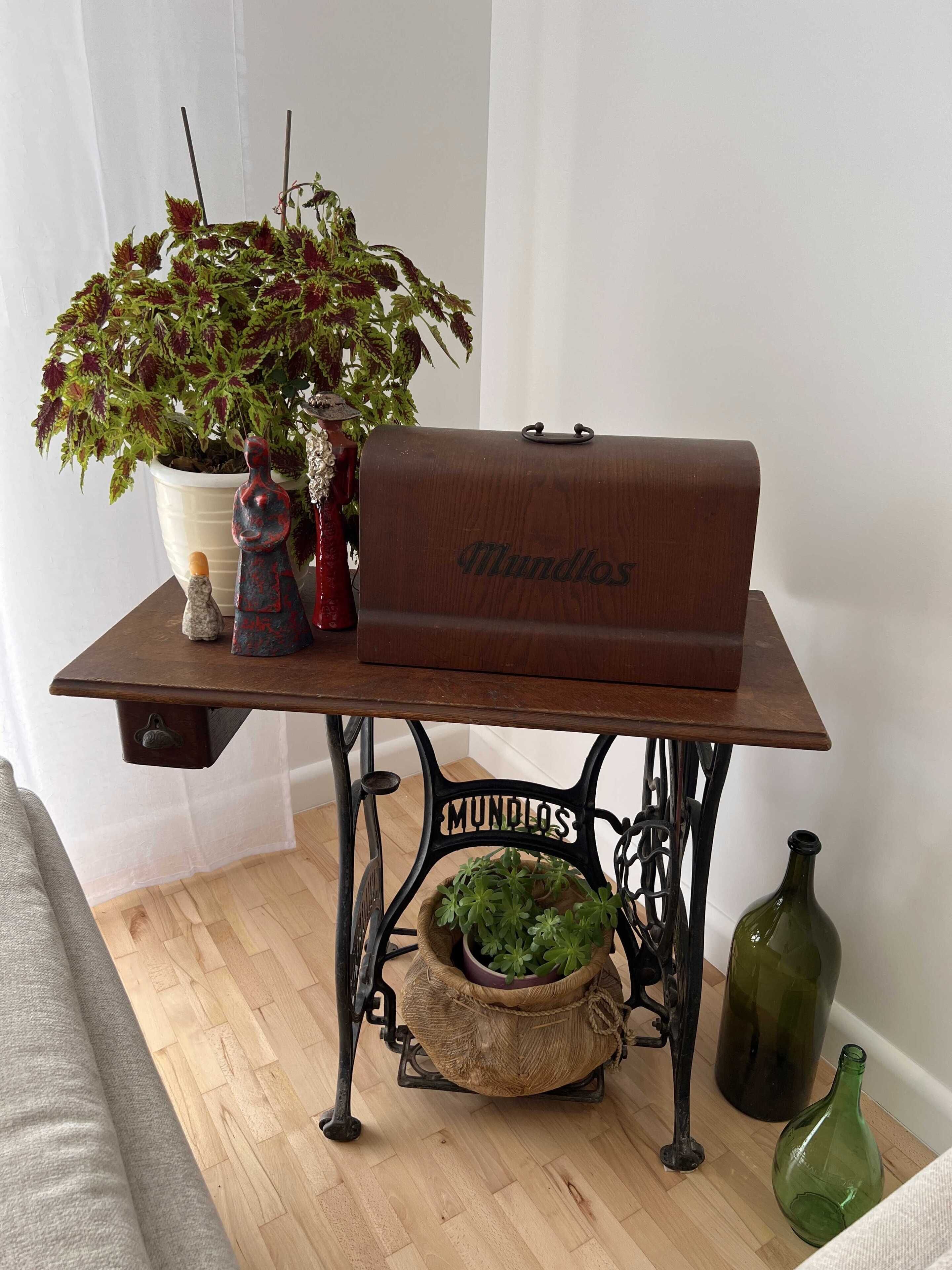 Uma mesa de madeira com uma máquina de costura antiga com a etiqueta 'MUNDOS', uma planta em vaso, uma figura decorativa e uma mala de época.