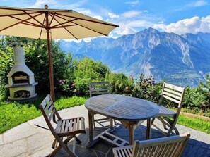 An outdoor patio with a round table and four chairs, a wooden pizza oven, and a large umbrella. - Cheechako by Interhome (Nendaz)