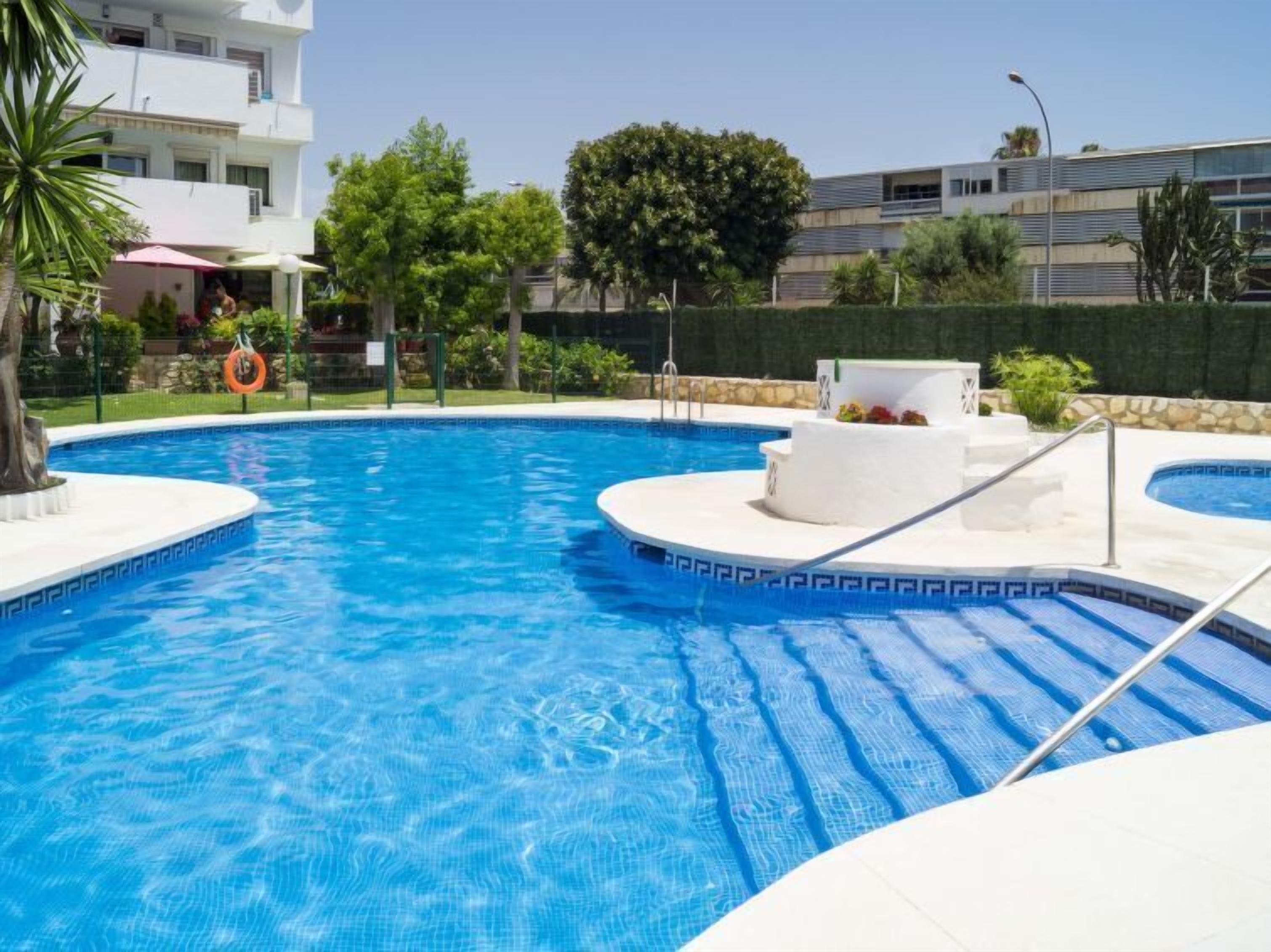 A swimming pool with a sun terrace, surrounded by greenery and a building in the background.