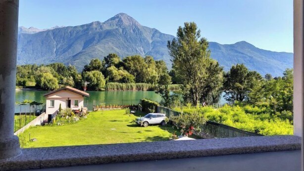 Blick auf einen See mit Bergen im Hintergrund, ein kleines Haus mit Rasen und ein geparktes Auto in der Nähe.