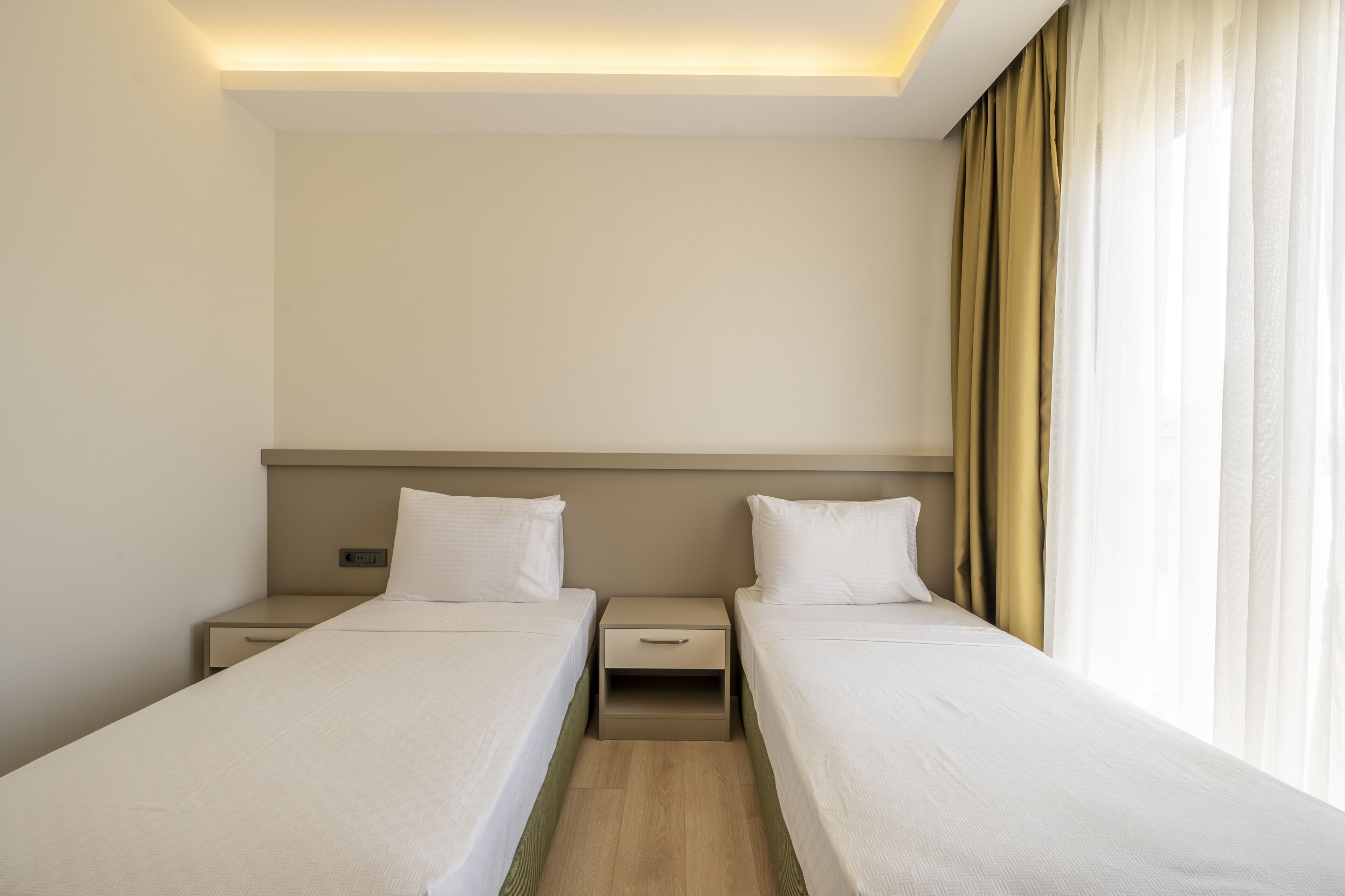 A hotel room with two single beds, a nightstand with a clock, and a window with curtains.