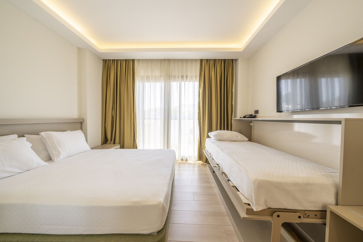 A hotel room with a large bed, a smaller bed, a TV, and a window with curtains.