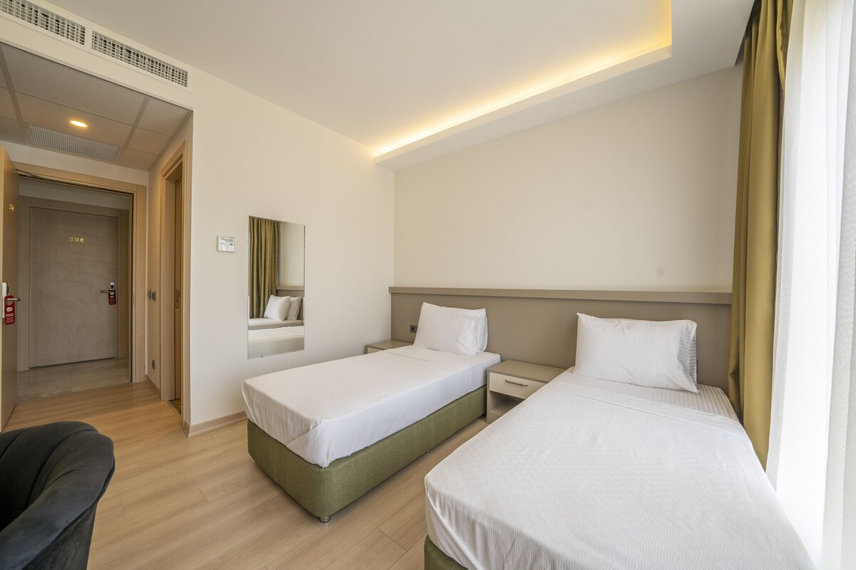 A hotel room with two single beds, a bedside table, a chair, a mirror, and a door leading to another room.
