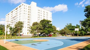 Ein Basketballplatz mit blau-orangenem Spielfeld, umgeben von Bäumen und einem weißen Gebäude mit zahlreichen Fenstern.