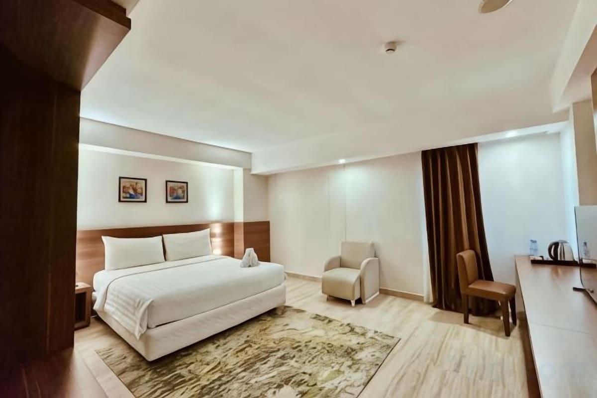 A hotel room with a bed, a desk, a chair, a nightstand, and a large window with curtains.