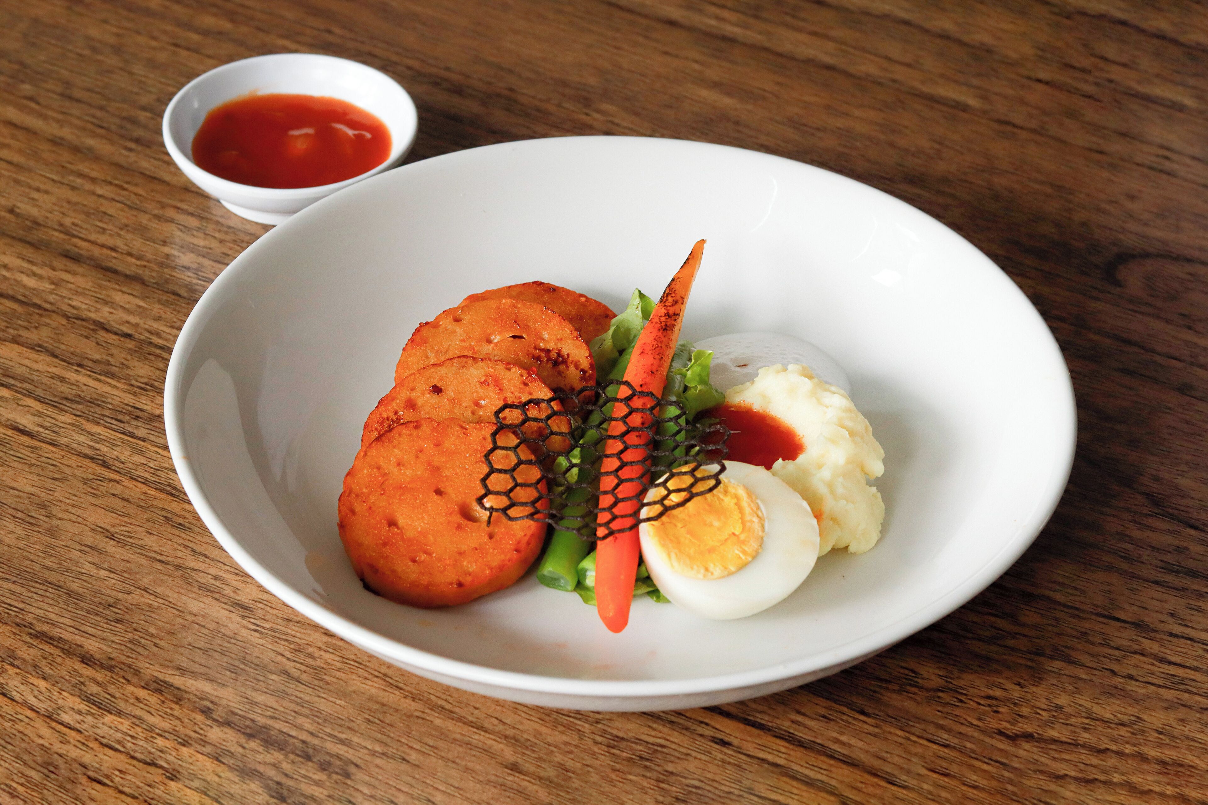 A white plate with food items including a boiled egg, mashed potatoes, and fried items, accompanied by a small bowl of sauce.