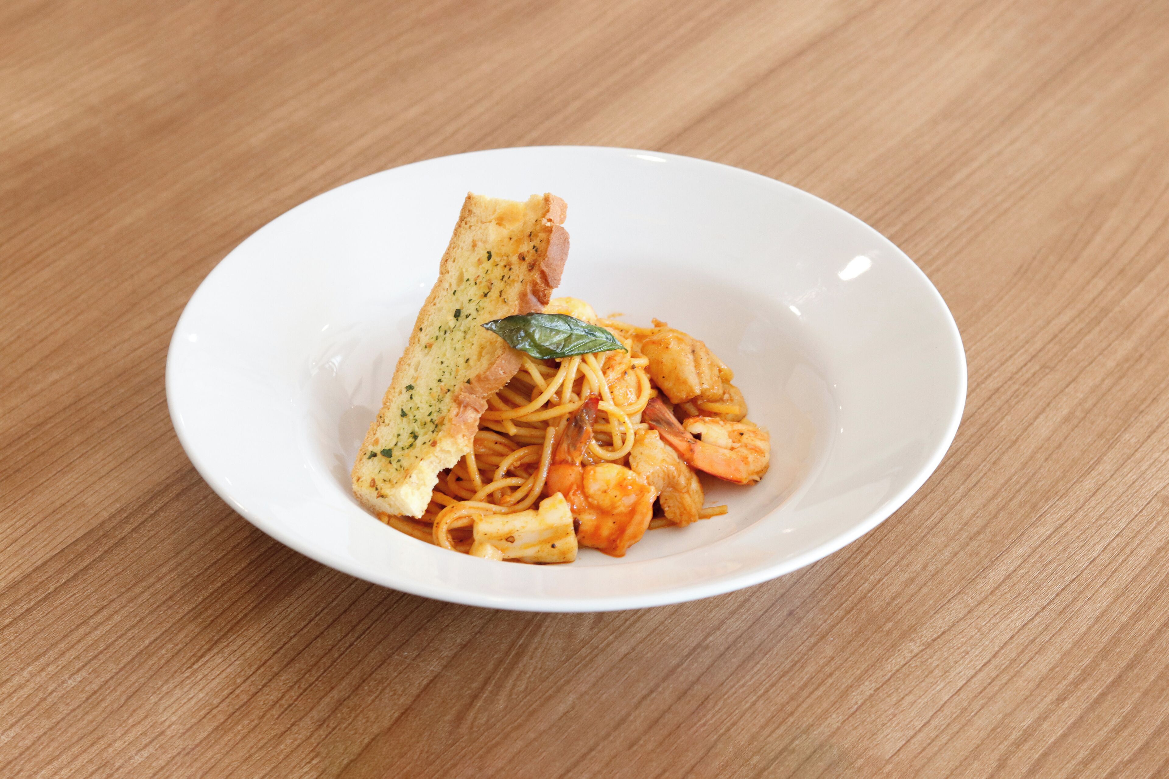 A white plate with spaghetti, shrimp, and a piece of bread on a wooden table.
