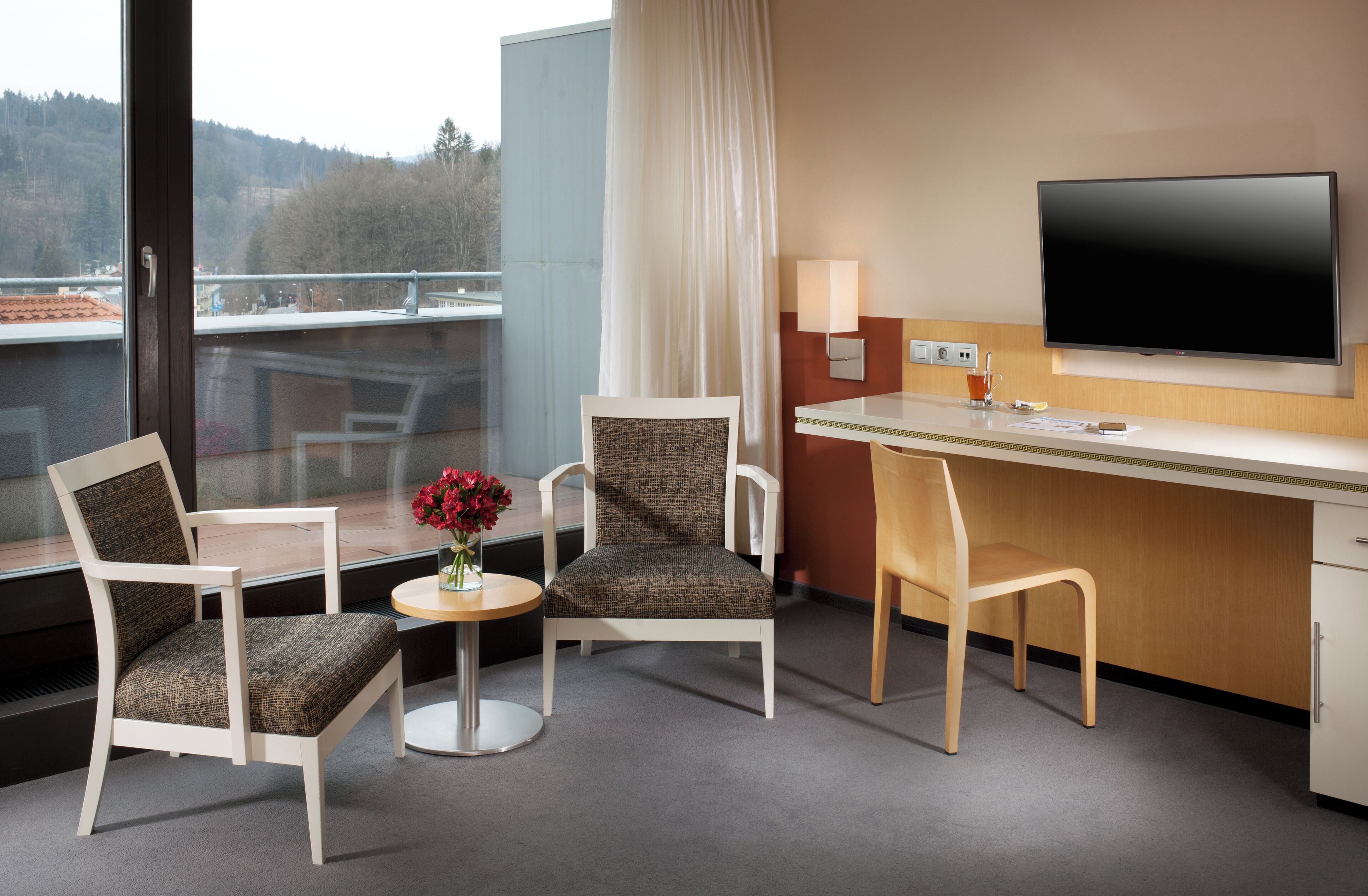 A hotel room with two chairs, a small table with a vase of flowers, a desk with a flat-screen TV, and a window offering a view of the outdoors.