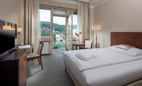 A hotel room with a bed, a desk with a TV, a chair, a table with flowers, and a view of a city and forest through the window.