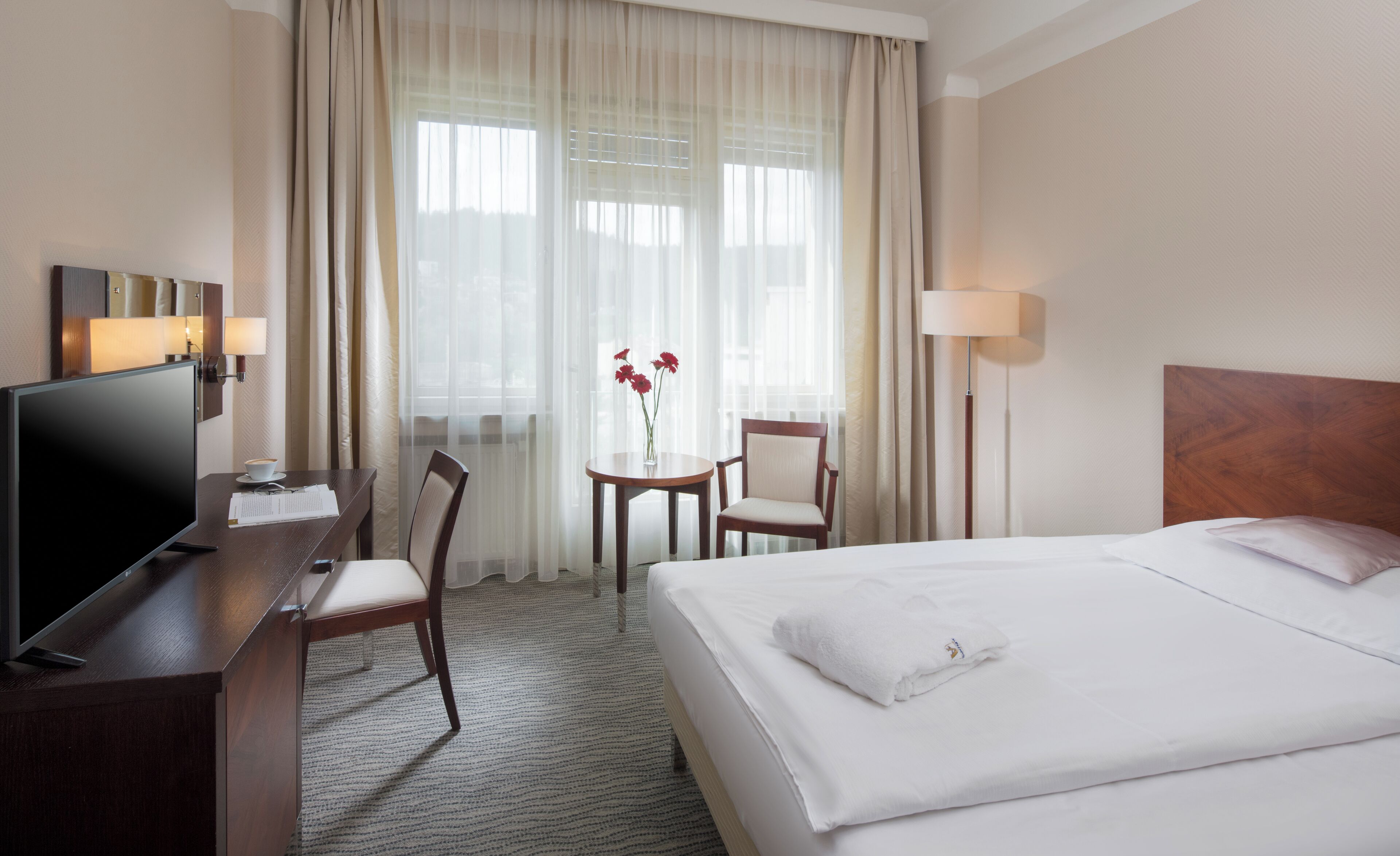 Habitación de hotel con cama, escritorio con televisor, silla, una mesita con flores y lámpara.
