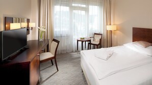 A hotel room with a bed, a desk with a TV, a chair, a small table with a lamp, and sheer curtains. - ALEXANDRIA Spa & Wellness hotel (Luhacovice)
