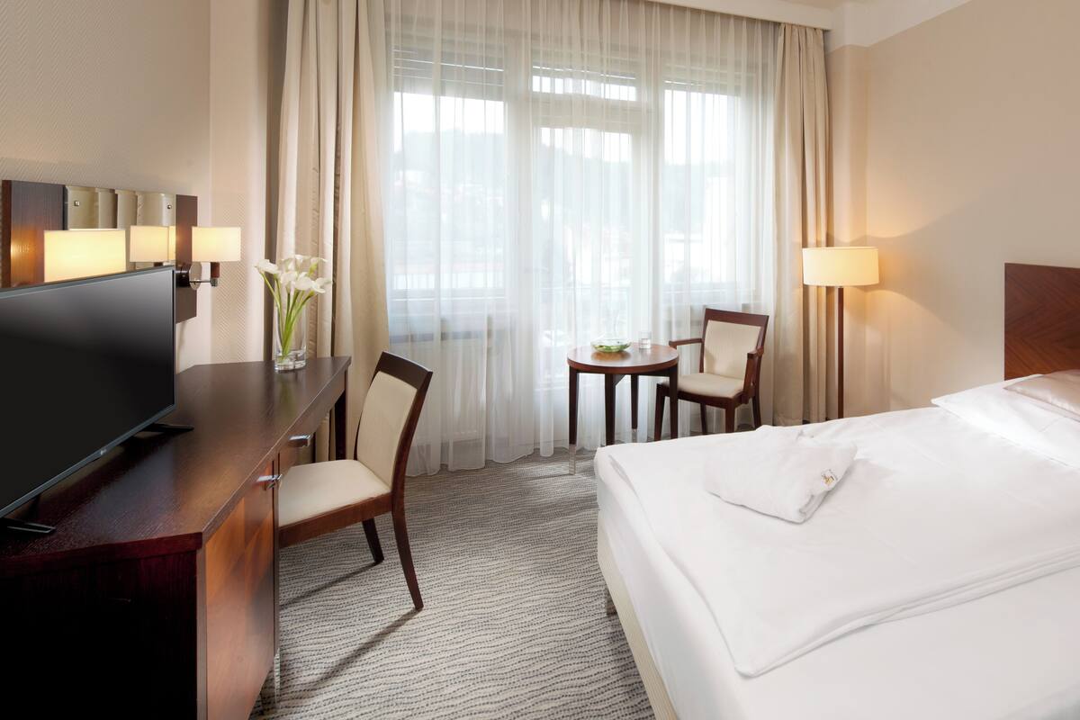 A hotel room with a bed, a desk with a TV, a chair, a small table with a lamp, and sheer curtains.