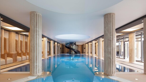 A spacious indoor pool with a central spiral staircase, surrounded by marble columns and glass doors. - ALEXANDRIA Spa & Wellness hotel (Luhacovice)