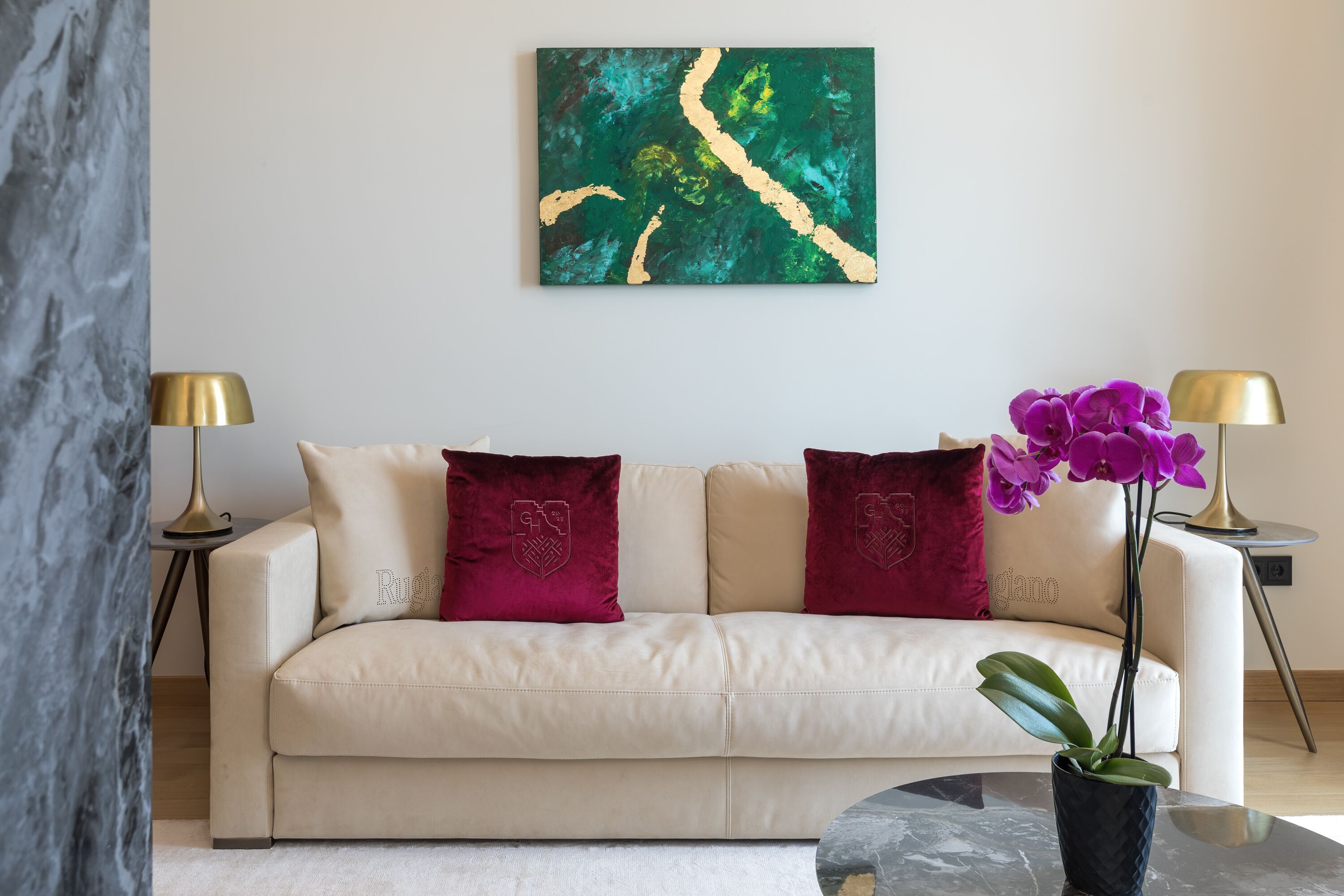 A modern living room with a beige sofa, two decorative pillows, a large abstract painting, a potted orchid, and two floor lamps.