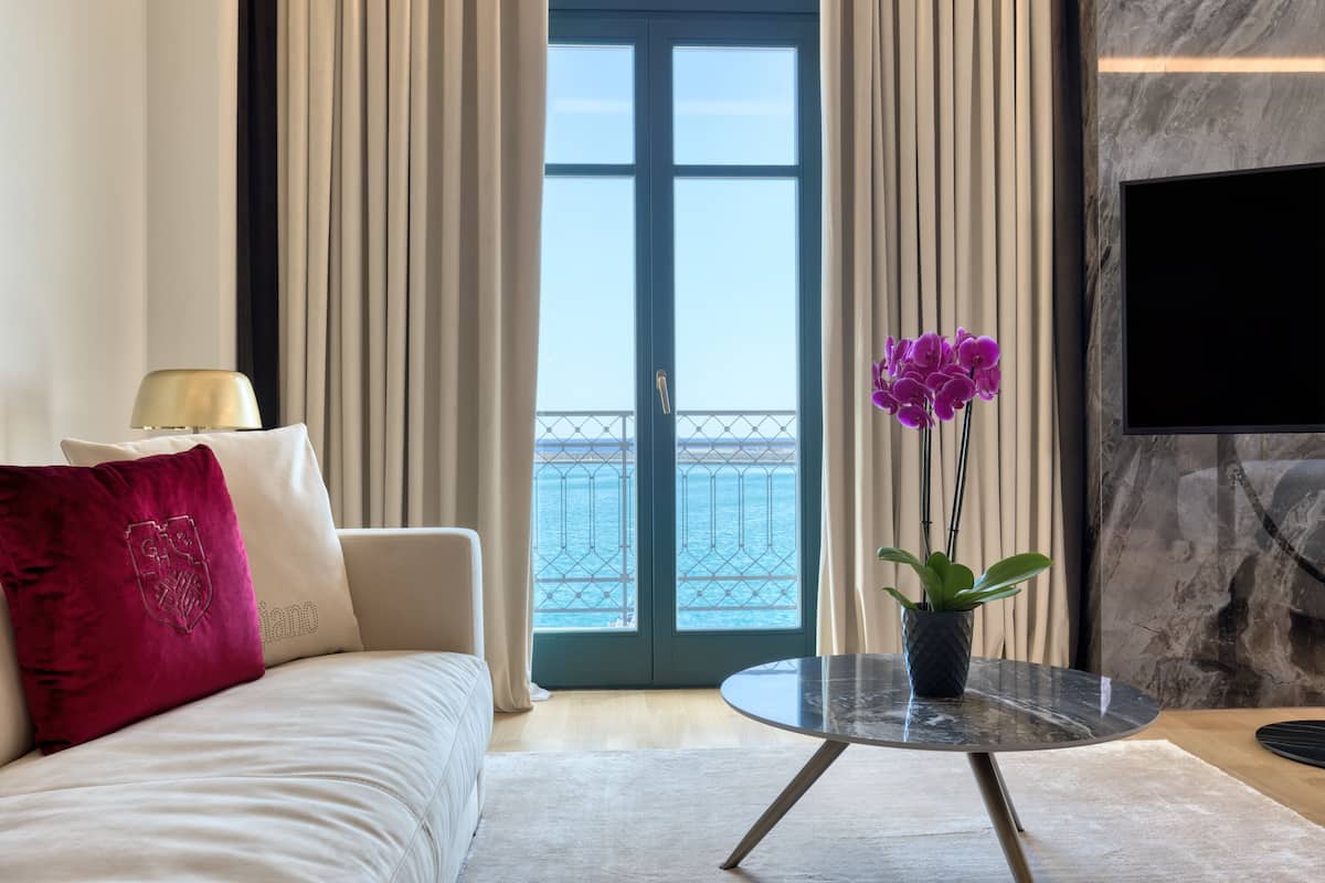 A hotel room with a sofa, a small table with a vase of flowers, a TV mounted on the wall, and a view of the sea through a large window.