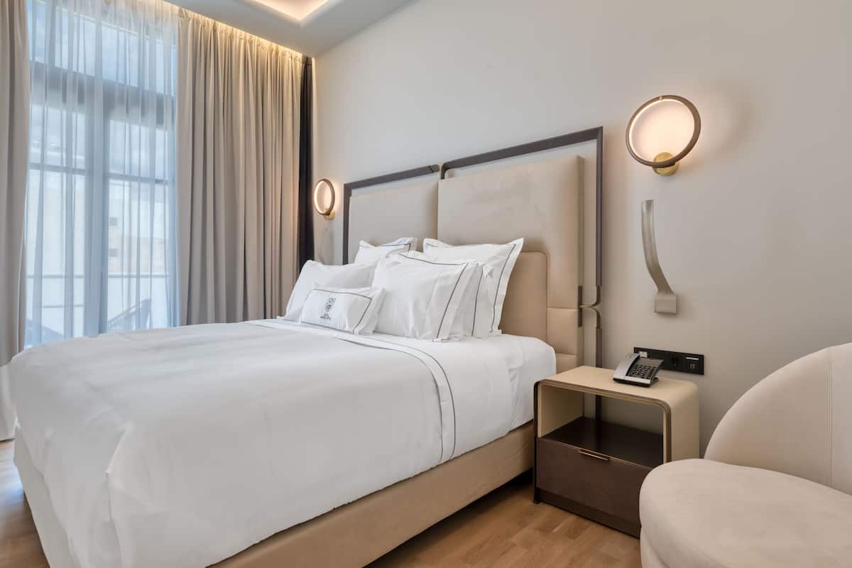 A modern hotel room with a large bed, bedside tables, and a view of the city through sheer curtains.