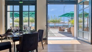 An indoor dining area with tables set for a meal, large windows, and outdoor umbrellas. - Grand Hotel Kalamata (Kalamata)