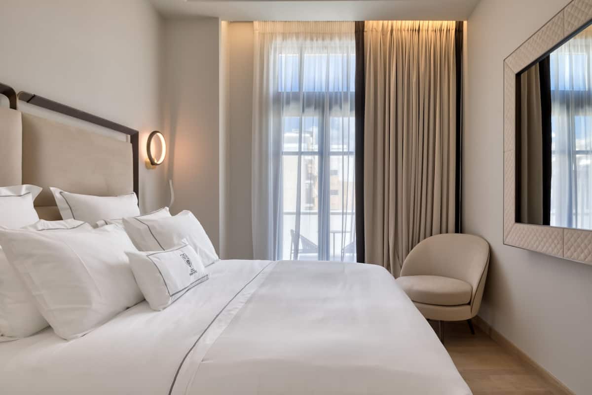 A modern hotel room with a large bed, a sitting area, and a view of the city through large windows.