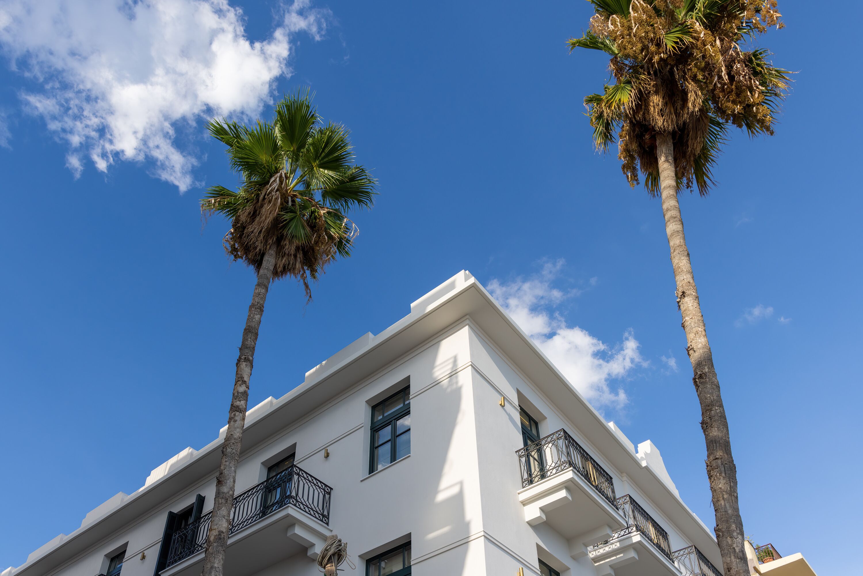 A white building with balconies and palm trees against a clear blue sky.