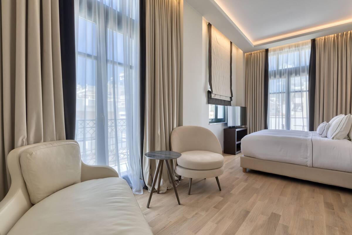 A modern hotel room with a large bed, a sofa, a small round table, and a chair. The room has large windows with curtains, a flat-screen TV, and a wooden floor.