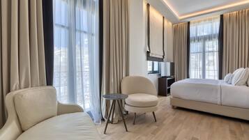 Ein modernes Hotelzimmer mit einem großen Bett, einer Couch, einem kleinen runden Tisch und einem Sessel. Das Zimmer verfügt über große Fenster mit Vorhängen, einen Flachbildfernseher und einen Holzboden.