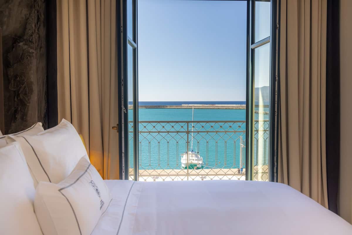 A hotel room with a large bed, white bedding, and a view of the sea through a balcony with a metal railing.