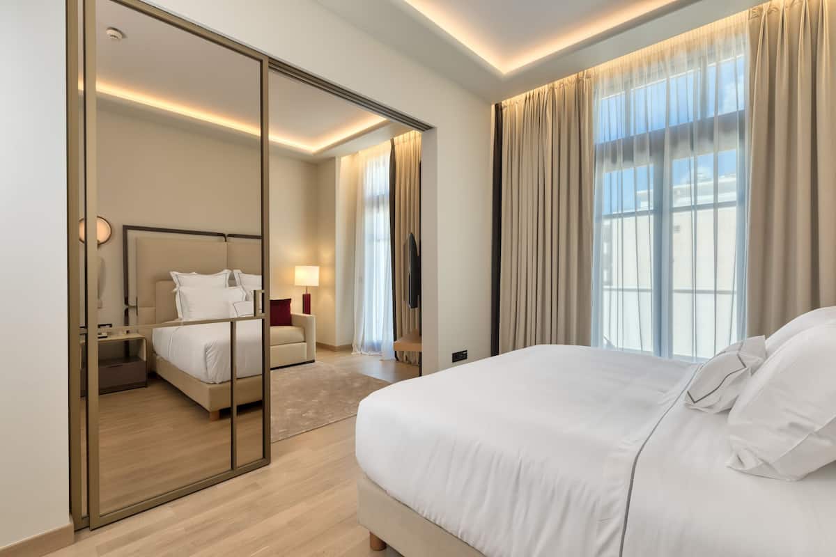 A modern hotel room with a large bed, a separate seating area, a mirror, and a view of the outdoors.