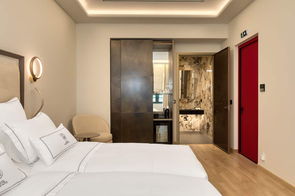 A hotel room with a bed, a chair, a desk, a minibar, and a bathroom visible through an open door.