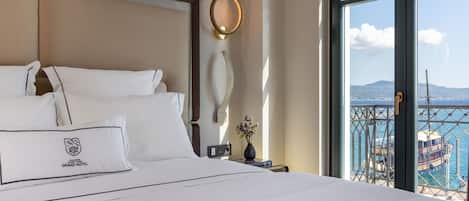 A hotel room with a large bed, a nightstand with a vase of flowers, a wall-mounted light fixture, and a view of a boat through a window.