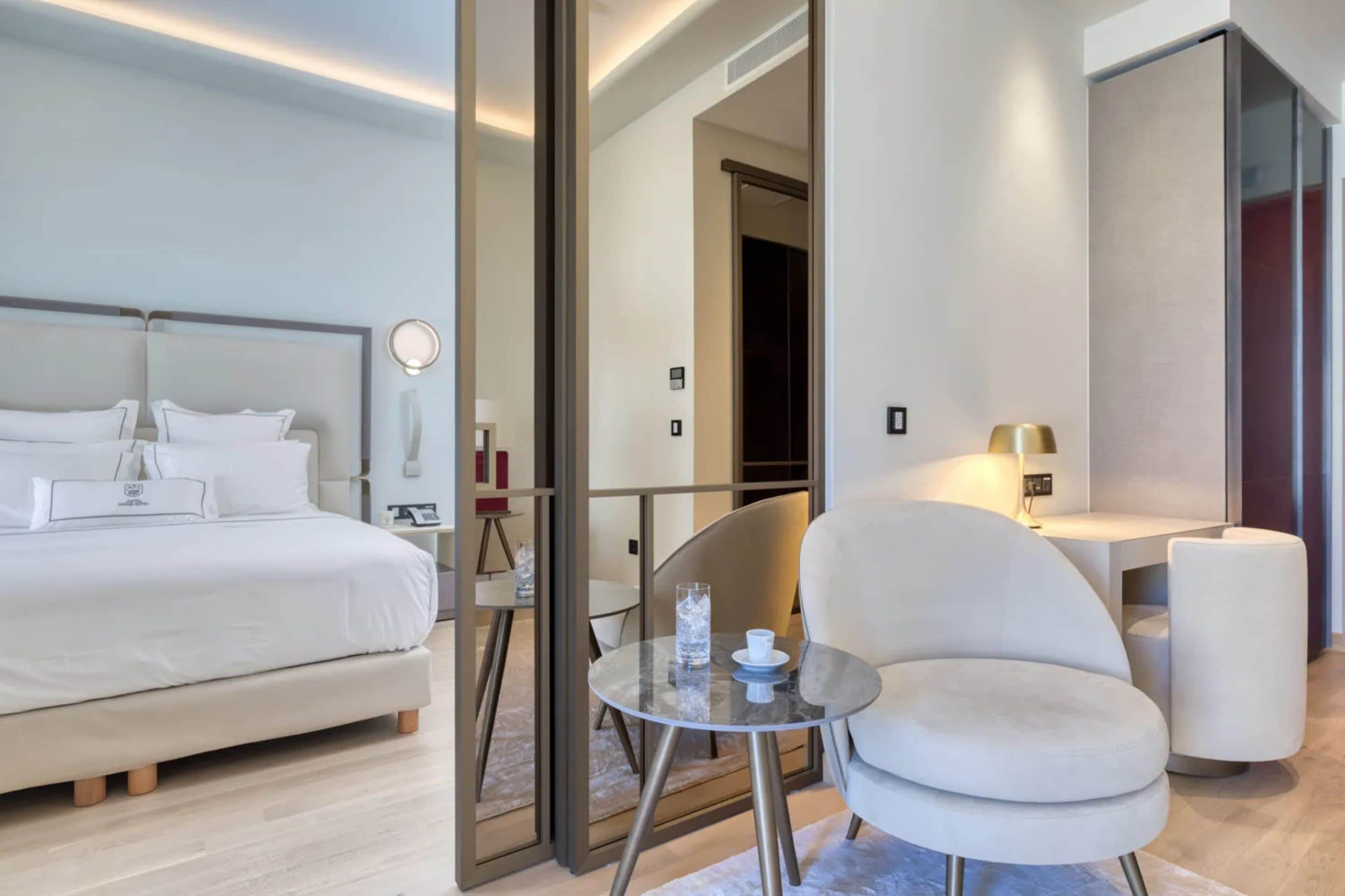 A modern hotel room with a large bed, a round table with a glass top, a white armchair, and a desk with a lamp.