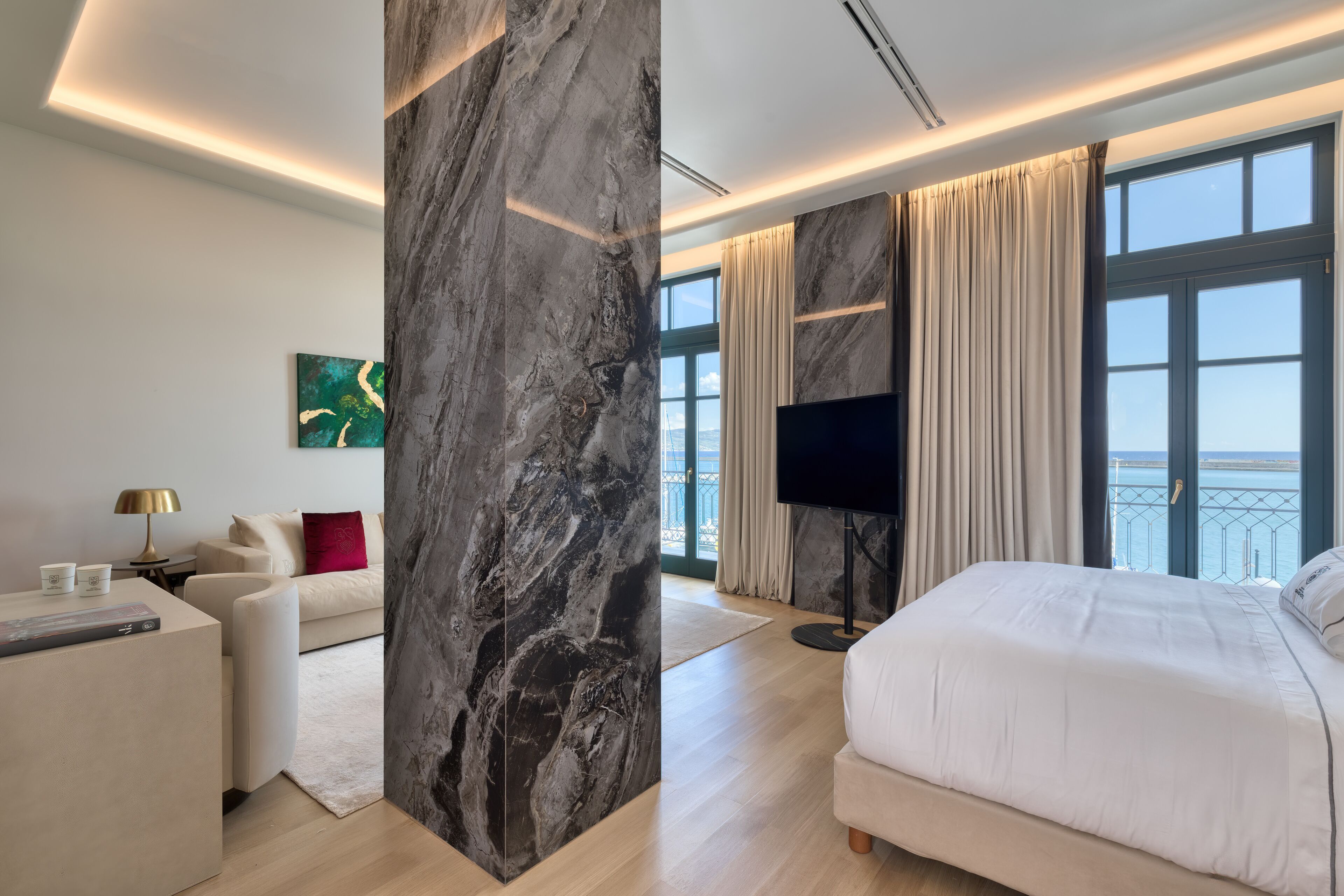 A modern hotel room with a large marble column, a bed, a TV, and a view of the sea.