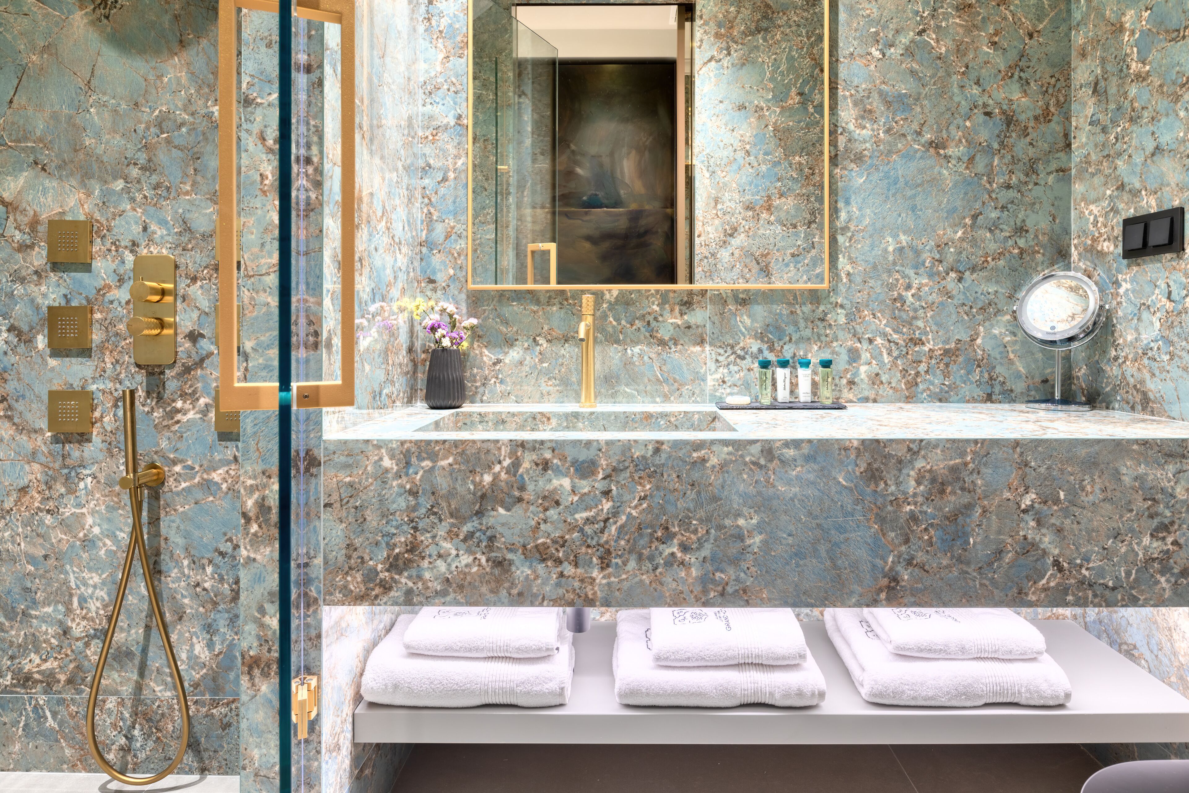A bathroom with a marble wall, a gold shower, a sink with toiletries, and towels on a shelf.