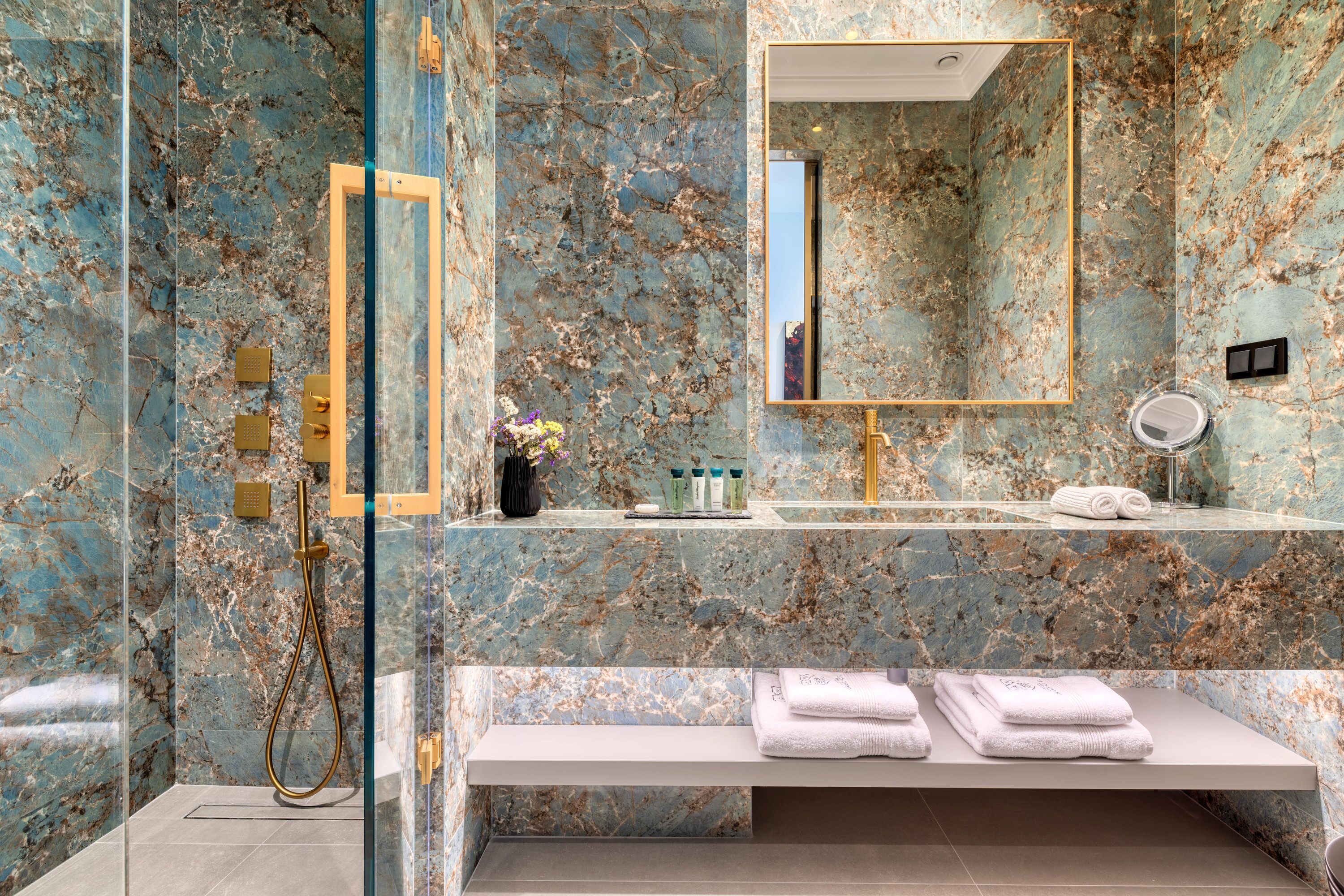 Un baño con pared de mármol, un espejo grande, un lavamanos y un banco con toallas.