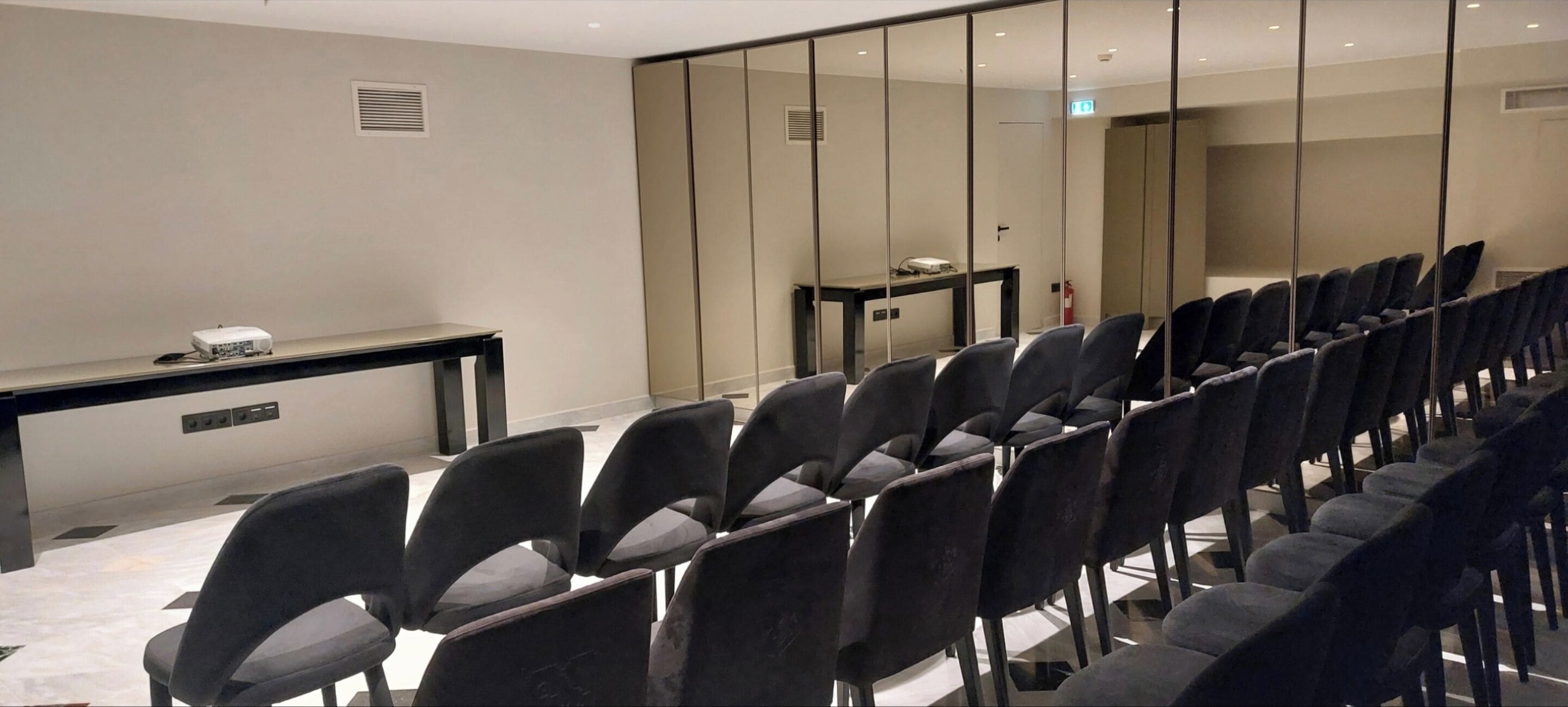 A conference room with rows of chairs, a projector on a table, and a large mirror wall.