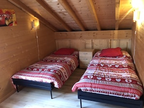A small, cozy room with two single beds, each with a red pillow and a patterned blanket. There is a small wooden nightstand with drawers between the beds. The room has wooden walls and a sloped ceiling with exposed beams.
